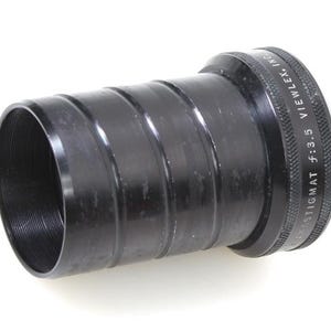 May include: A black camera lens with a ridged body and threaded interior. The lens has the text "STIGMAT f:3.5 VIEWLEX, INC." engraved on the outer ring. The lens is cylindrical and appears to be made of metal.