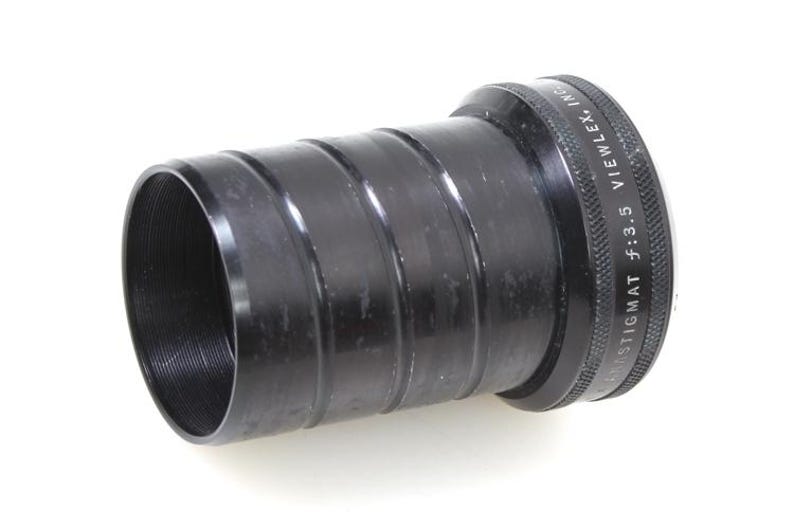 May include: A black camera lens with a ridged body and threaded interior. The lens has the text "STIGMAT f:3.5 VIEWLEX, INC." engraved on the outer ring. The lens is cylindrical and appears to be made of metal.