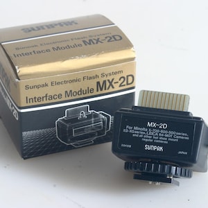 May include: A Sunpak Electronic Flash System Interface Module MX-2D, with its original box. The box is gold and black, with the Sunpak logo and product information. The module is black with a gold connector and the text "MX-2D".