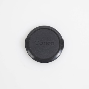 May include: A black Canon lens cap, circular in shape, with the brand name embossed in the center. The cap is made of a matte material and is set against a plain white background.