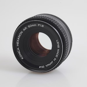 May include: A Konica Hexanon AR 50mm F1.8 camera lens. The lens is black with silver text, including "Lens Made in Japan 55ø". The lens has a ribbed focus ring and a clear glass element.