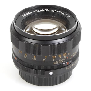 May include: A black Konica Hexanon AR 57mm F1.4 camera lens. The lens has a series of measurement markings and numbers, including f/1.4, and a focus ring. The lens is designed for use with cameras.