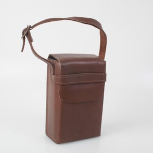 Op de afbeelding: Een bruine leren etui met een handvat en een klepsluiting. De etui heeft een rechthoekige vorm en een riem die om de behuizing gaat. De handgreep is aan de bovenkant van de etui bevestigd. De etui is gemaakt van glad, glanzend leer.