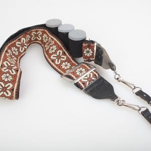May include: A camera strap with a floral pattern in brown, cream, and black. The strap has black leather ends and silver-colored metal clips. Three black spools with gray lids are also present.