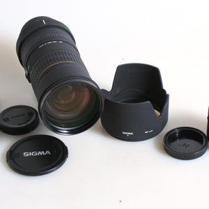 Puede incluir: Un objetivo Sigma APO 50-500mm F4-6.3EX negro con parasol, tapa de objetivo y teleconvertidor. El objetivo está sobre una superficie blanca.