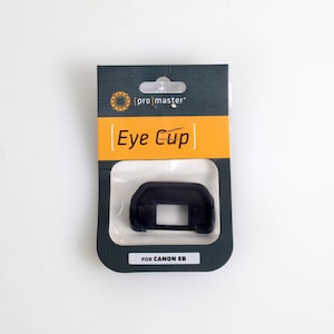 May include: A black eye cup for a Canon EB camera, packaged in a dark green and orange card. The package has the text "Eye Cup" and "FOR CANON EB". The eye cup is a camera accessory.