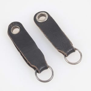 Könnte beinhalten: Zwei schwarze Leder-Schlüsselanhänger mit silbernen Metall-Schlüsselringen. Jeder Schlüsselanhänger hat oben eine Metallöse. Das Leder weist Gebrauchsspuren auf. Auf weißem Hintergrund.