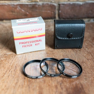 May include: A set of three black camera lens filters in a black leather case with a snap closure. The filters are labelled "Quantaray Professional Filter Kit".