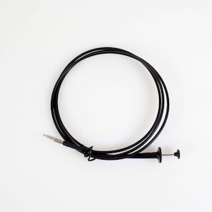 May include: A black mechanical shutter release cable featuring a flexible coiled cord, a metallic threaded tip at one end, and a plunger-style button mechanism at the other end, designed for remote camera operation and long exposure photography.