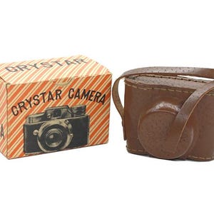 May include: A vintage camera and case set. The camera box is orange and white striped with the words "CRYSTAR CAMERA" and a camera illustration. The brown leather camera case has a strap.