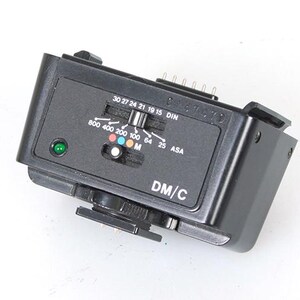 May include: Black camera flash unit with a control panel featuring ASA and DIN settings, along with a green indicator light. The unit has a sliding switch and the text "DM/C" is visible.