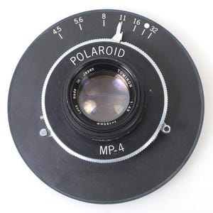 May include: A black Polaroid MP-4 camera lens assembly. The lens is surrounded by a black circular frame with white markings for aperture settings. The lens itself is labeled "TOMINON" and "Made in Japan".