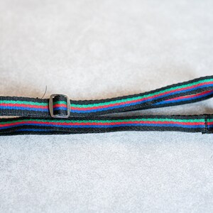 Puede incluir: Una correa de cámara negra con rayas rojas, azules y verdes. La correa tiene una hebilla de metal.
