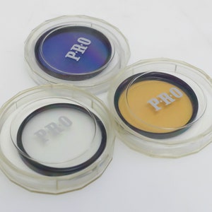 May include: Three clear plastic containers with black lids, each containing a round filter with the word "PRO" printed on it. The filters are blue, yellow, and white.