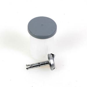 Può includere: Un contenitore di plastica trasparente con un coperchio grigio e un componente metallico argento e grigio. Il contenitore è cilindrico e il coperchio è piatto. Il componente metallico ha una forma complessa.