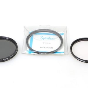 May include: Three 72mm camera lens filters. A black filter with the text "Hoya POLARIZER 72mm" printed on it. A black filter with the text "QUANTA RAY 72mm 1A" printed on it. A black filter with the text "SPRINTONE 72MM DIFFUSER" printed on it.