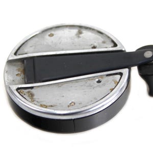 Puede incluir: Un objeto metálico circular con un asa negra. El objeto tiene una parte superior plateada con una banda negra alrededor del borde. El asa es negra y está sujeta a la parte superior con un pequeño pasador metálico.