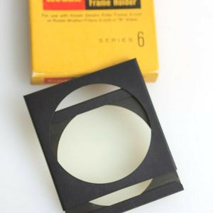 Puede incluir: Un soporte de marco de filtro de gelatina negro con un filtro blanco, junto a una caja amarilla Kodak. La caja dice "Kodak Gelatin Filter Frame Holder Series 6". El soporte del marco es para filtros de gelatina Kodak.