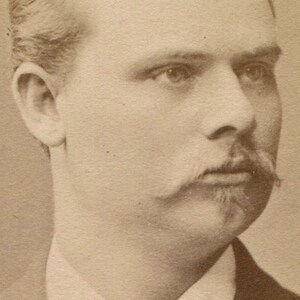 May include: Sepia-toned portrait of a man with light hair, a mustache, and a goatee. He is wearing a suit jacket and a collared shirt with a tie. The photograph is a close-up of the man's face and upper body.