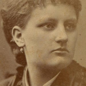 May include: Sepia-toned portrait of a woman with short, wavy hair. She wears a dark garment with a decorative collar and a small brooch. The image is a vintage photograph, likely from the late 19th century.