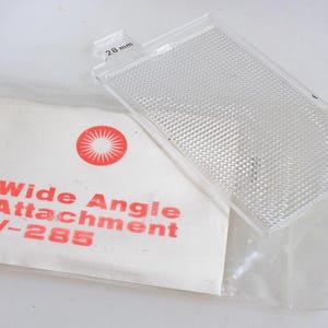Peut inclure: Un accessoire grand angle rectangulaire transparent avec une surface texturée, à côté d'un paquet blanc. Le paquet porte le texte rouge "Wide Angle Attachment V-285" et un logo rouge en forme d'éclat de soleil. L'accessoire mesure 28 mm.