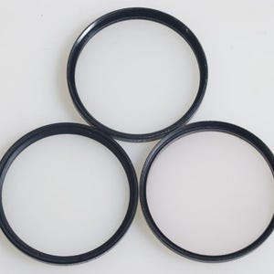 May include: Three circular black camera lens filters on a white background. Each filter has a black rim and a clear centre. The filters are of different sizes and are arranged in a triangular pattern.