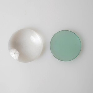 May include: Two round objects on a white surface. One is a translucent, white, dome-shaped object with a starburst pattern at the base. The other is a flat, circular, light green object with a dark green border.