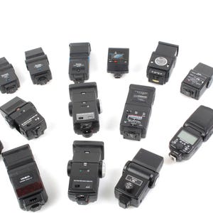 May include: A collection of 14 black flash units for cameras. The flash units are arranged in a circle on a white background.