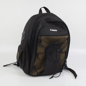 May include: Black and olive green Canon backpack with multiple compartments and adjustable straps. Features the Canon logo in white on the top. Includes a top handle and side pockets for storage.