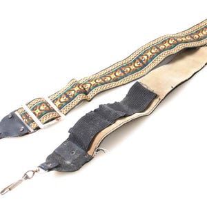 May include: A guitar strap with a patterned design in shades of green, orange, and tan. The strap has a black leather end with a metal clip. The other end is a light tan suede with a black fabric sleeve.