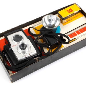 May include: A black box containing a silver and black camera, a flash, and a roll of Kodakcolor film. The box is labelled "Brownie Starflex Outfit".