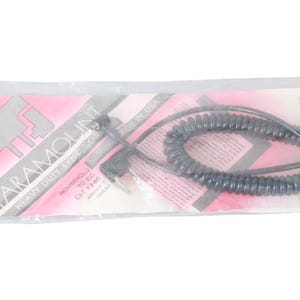 Könnte beinhalten: Ein schwarzes Spiralsynchronisationskabel, verpackt in einer transparenten Plastiktüte. Die Verpackung trägt den Text "PARAMOUNT HEAVY DUTY SYNC CORD" und ein rosa-weißes Design. Das Kabel ist mit "MADE IN USA" gekennzeichnet.