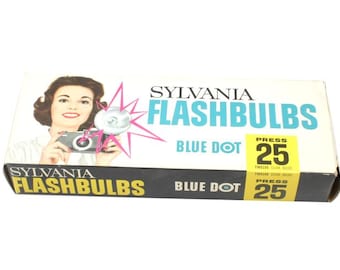 May include: Vintage Sylvania Flashbulbs box. The box features a woman holding a camera with a flash. The text on the box reads "SYLVANIA FLASHBULBS BLUE DOT" and "PRESS 25". The box is white, black, and blue.