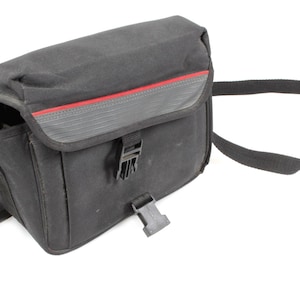 May include: Black camera bag with a red accent stripe. The bag has a flap closure with a buckle and a black shoulder strap. The bag is made of a durable, water-resistant material.