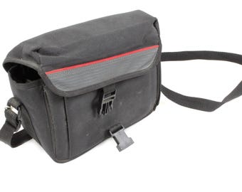 May include: Black camera bag with a red stripe detail. The bag features a flap closure with a buckle and a black shoulder strap. Constructed from a durable, water-resistant material.