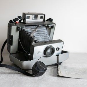 Puede incluir: Una cámara Polaroid Automatic 320 vintage, plateada y negra, con una lente negra y una correa negra. La cámara está abierta, revelando el fuelle. Una tapa de lente negra con la palabra "POLAROID" está al lado de la cámara.
