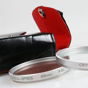 Puede incluir: Un filtro de lente de cámara plateado con una funda de cuero negro y un forro de terciopelo rojo. El filtro está etiquetado como "CRYSTAL-OPTICS F-DL 49mm".