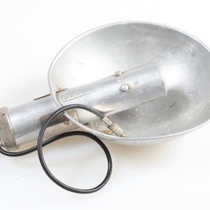 May include: Vintage silver-toned camera flash unit with a large, rounded reflector. The flash has a cylindrical body with a textured grip and a black cord. The unit is marked with the letters "P-Z-N" on the side.