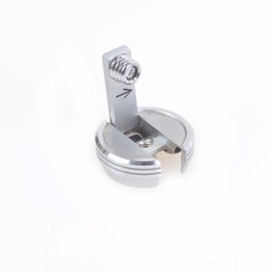 Puede incluir: Un pequeño componente metálico de color plateado con un tornillo y una flecha. El componente tiene una base circular con un diseño acanalado y una pieza rectangular fijada en ángulo. El fondo es blanco liso.