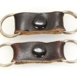 May include: Two dark brown leather key fobs with silver key rings. Each fob features a black button. The leather shows signs of wear, with visible stitching and a slightly distressed appearance. The key rings are circular.
