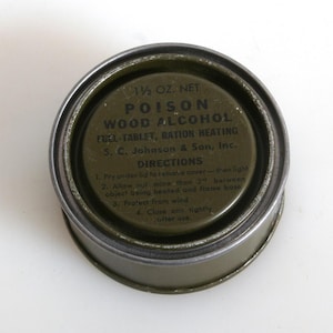 May include: A vintage, olive-drab metal can with a silver rim. The lid features printed text: "POISON WOOD ALCOHOL FUEL-TABLET, RATION HEATING." Includes directions and the manufacturer's name, S. C. Johnson & Son, Inc. The can contains 1 1/2 oz.