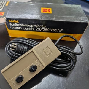 May include: A vintage Kodak audioviewer/projector remote control 210/260/260AF. The beige remote has two black buttons and a black cord. The box is black and yellow with the Kodak logo.