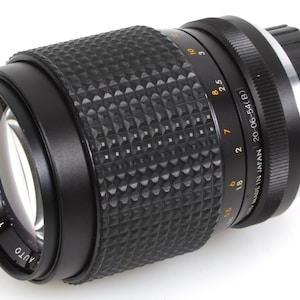 May include: Black camera lens with a textured grip. The lens has a focal length of f=135mm and an aperture of 1:2. The lens is marked "AUTO" and "MADE IN JAPAN".