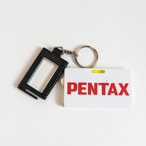 May include: A black keychain with a clear plastic window and a silver key ring. The keychain is attached to a white card with the word "PENTAX" printed in red letters.