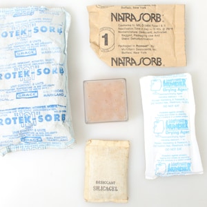 Op de afbeelding: Een verzameling van zes droogzakjes, waaronder een doorzichtig plastic zakje met een roze gel, een wit zakje met blauwe tekst dat "Laminer Drying Agent" zegt, een wit zakje met blauwe tekst dat "Desiccant Silicagel" zegt, en drie bruine papieren zakjes met de tekst "NATRASORB" en "PROTEK SORB".