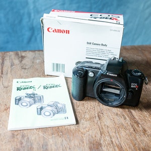May include: A black Canon EOS Rebel XS camera with a lens attached. The camera is sitting on a wooden table next to its instruction manual and original box. The box is white with red and black text that reads "Canon EOS Rebel XS Still Camera Body".