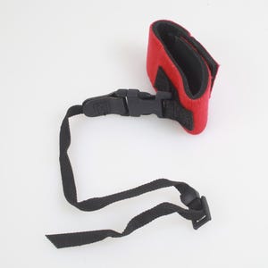 May include: A red padded strap with a black interior lining, featuring a black plastic side-release buckle attached to a long, adjustable black nylon webbing strap with a sliding plastic adjuster for secure fastening and length control.