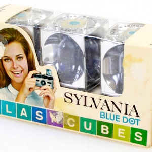 May include: A box of three Sylvania Blue Dot Flash Cubes. The box features a woman holding a camera and the text "FLASH CUBES" and "SYLVANIA BLUE DOT".