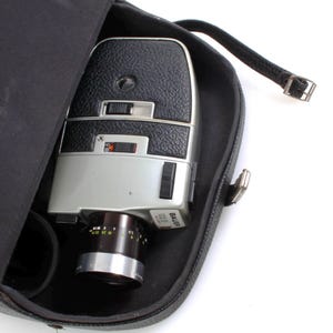 Pode incluir: Uma câmera de filme Super 8 Bauer prateada e preta em uma caixa de transporte preta. A câmera tem uma lente com uma anel de zoom e uma alça de couro preta.