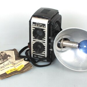 Puede incluir: Una cámara vintage en blanco y negro con un accesorio de flash. La cámara tiene una correa negra y el flash tiene una bombilla azul. La cámara tiene el texto "Miraflex" y "Six Twenty" en ella. La cámara está acompañada de un folleto titulado "Taking Good Pictures is Fun".